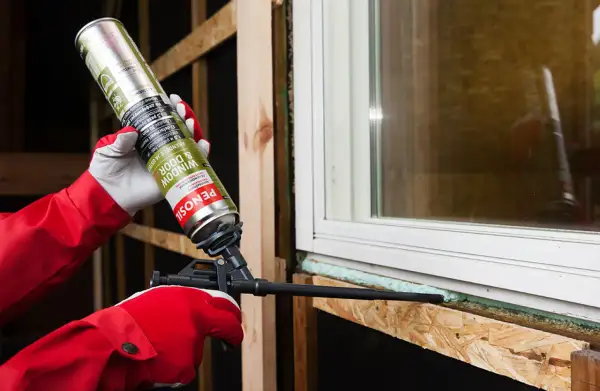 Persona con guantes rojos aplica espuma de poliuretano no expansiva alrededor de una ventana.