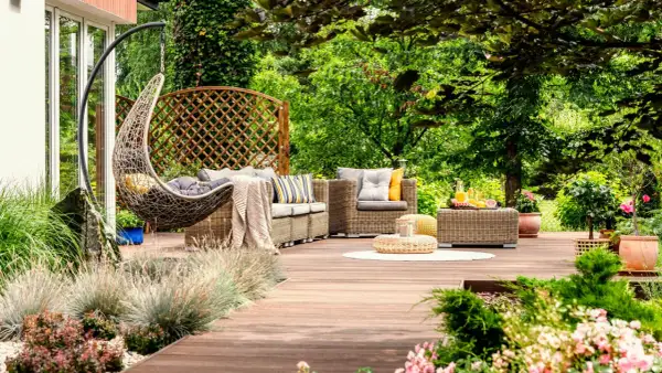 Un acogedor rincón en un jardín de verano, con muebles de mimbre, una silla colgante y vegetación exuberante.